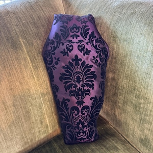 Può includere: Un cuscino a forma di bara con un motivo damascato viola e nero. Il cuscino è seduto su un divano di velluto verde.