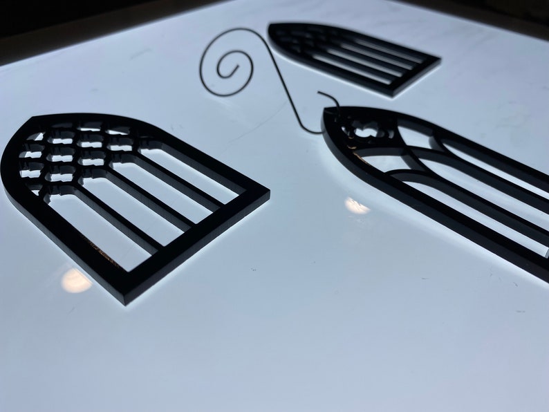 May include: Two black metal window ornaments with a decorative design. The ornaments are shaped like arched windows with a grid pattern.