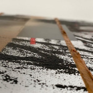 May include: Close-up of a textured, abstract art piece with a brown, wooden strip running down the center. The piece features black, white, and gray paint with a hint of red.