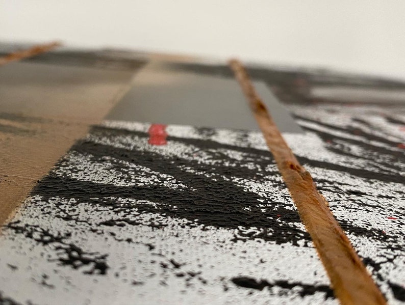 May include: Close-up of a textured, abstract art piece with a brown, wooden strip running down the center. The piece features black, white, and gray paint with a hint of red.