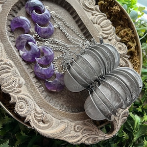 May include: A set of five amethyst crescent moon tea infusers with silver chains hanging from a wooden tray. The infusers are arranged in a semi-circle with the chains overlapping. The tray is decorated with a floral design.