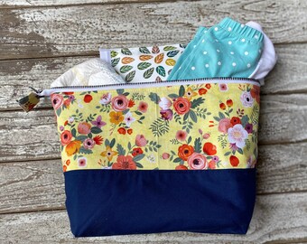 Puede incluir: Una bolsa de pañales con estampado floral con fondo amarillo y base azul marino. La bolsa está abierta, revelando un pañal, pantalones de bebé y un artículo con estampado de hojas. La bolsa tiene una cremallera blanca y una pequeña etiqueta floral.