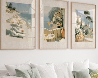 May include: Three framed landscape art prints depicting coastal scenes hang above a white sofa with green pillows. A wooden coffee table displays lemons and a vase of flowers. The room has a neutral color palette.