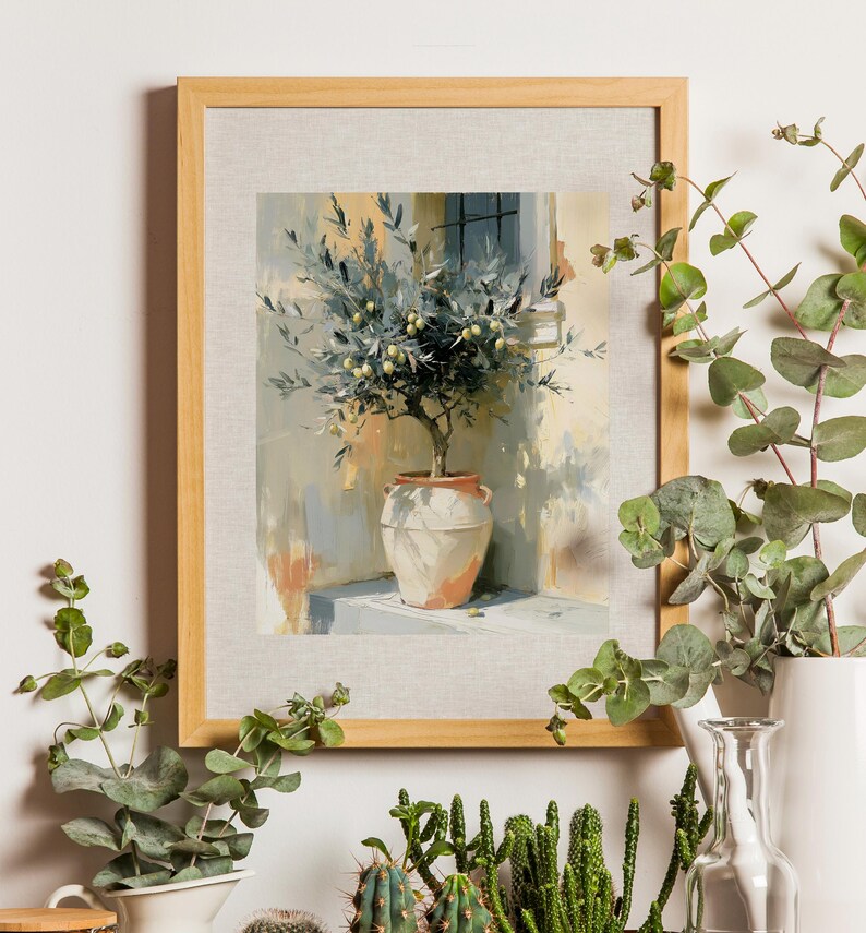 May include: A framed art print depicts an olive tree in a terracotta pot. The painting has a light color palette with a beige frame. The artwork is surrounded by greenery, including eucalyptus and cacti, creating a natural, decorative display.