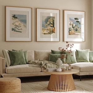 Set of 3 Italian coastal Amalfi Coast art prints in medium oak frames on a gallery wall above a wide beige sectional sofa with sage green velvet cushions — a bright earthy living room with bamboo blinds, natural wood round coffee table, and garden visible through large window