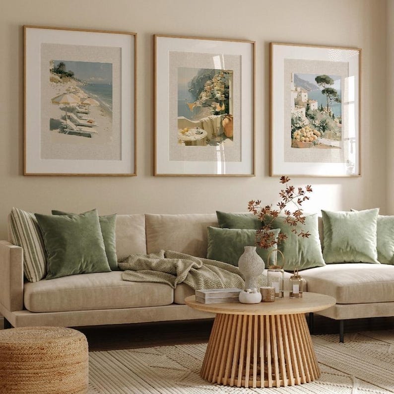 Set of 3 Italian coastal Amalfi Coast art prints in medium oak frames on a gallery wall above a wide beige sectional sofa with sage green velvet cushions — a bright earthy living room with bamboo blinds, natural wood round coffee table, and garden visible through large window