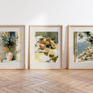 May include: Three framed art prints featuring watercolor paintings. One depicts a potted olive tree, another a still life of lemons, and the third a coastal scene with a window view. The frames are light-colored wood, and the artwork has a soft, warm color palette.