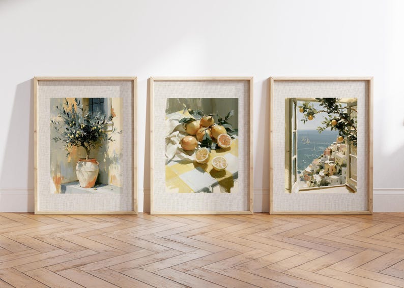 May include: Three framed art prints featuring watercolor paintings. One depicts a potted olive tree, another a still life of lemons, and the third a coastal scene with a window view. The frames are light-colored wood, and the artwork has a soft, warm color palette.