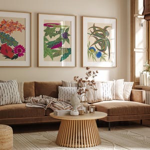 May include: Three framed botanical art prints with colorful floral designs hang above a brown sectional sofa with striped pillows and a throw blanket. A round wooden coffee table and a woven ottoman complete the living room setting.