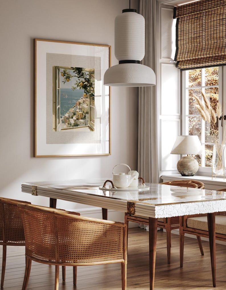 May include: A dining room scene featuring a rectangular table with a white and gold patterned top, surrounded by woven rattan chairs. A framed artwork depicts a coastal view. A white pendant light hangs above the table, and a window with a bamboo shade is visible.