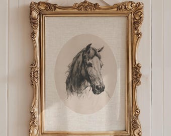 May include: A rectangular gold ornate frame containing a charcoal-style drawing of a horse head inside an oval border. The frame is topped with a white satin ribbon bow. The artwork is set against a light-colored, textured fabric background.