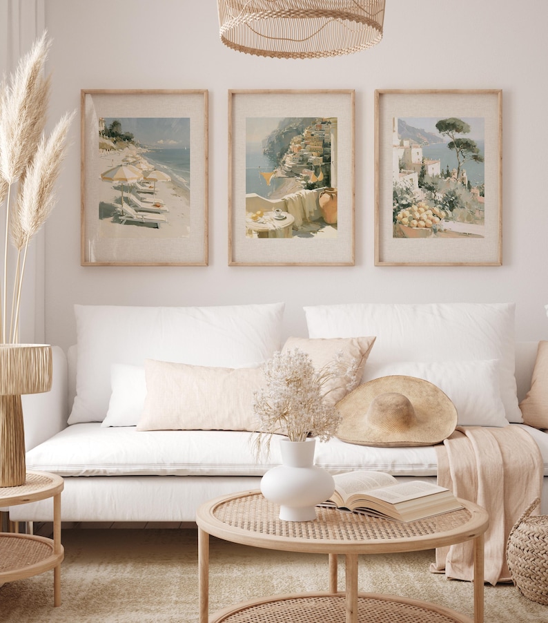Three Amalfi Coast Italian coastal art prints in matching natural wood frames hung as gallery wall above white linen sofa in a bright boho living room — beach umbrellas, Positano hillside, and Amalfi lemon terrace prints with woven rattan pendant light and pampas grass decoration