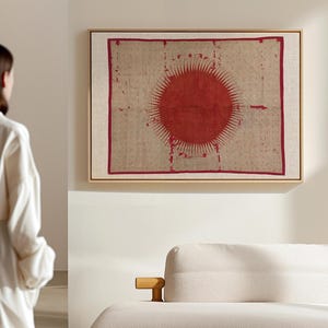 Peut inclure: Une œuvre d'art murale rectangulaire encadrée présentant un fond en tissu beige texturé avec un grand motif de soleil rouge vif au centre. Le design du soleil comprend des rayons rayonnants et est entouré d'une fine bordure rouge, le tout dans un cadre en bois clair.