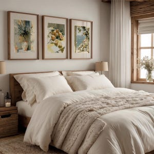 May include: A bedroom scene featuring a wooden bed frame with white bedding and a beige cable-knit throw blanket. Above the bed are three framed watercolor prints with botanical and coastal themes. A nightstand and window are visible.