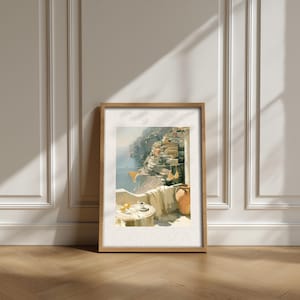 Single Positano balcony view Italian coastal oil painting print in natural light oak frame leaning against a white classical panelled wall on a herringbone parquet wood floor — terracotta amphora jug, linen cloth throw, espresso coffee cup and lemons on a marble table with Positano hillside in background