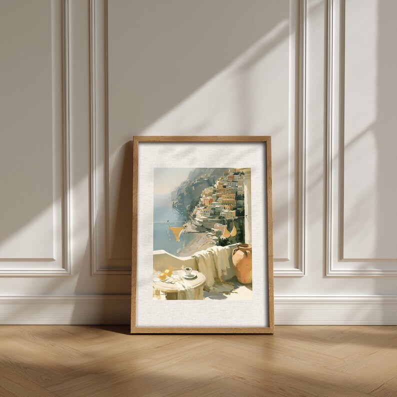 Single Positano balcony view Italian coastal oil painting print in natural light oak frame leaning against a white classical panelled wall on a herringbone parquet wood floor — terracotta amphora jug, linen cloth throw, espresso coffee cup and lemons on a marble table with Positano hillside in background