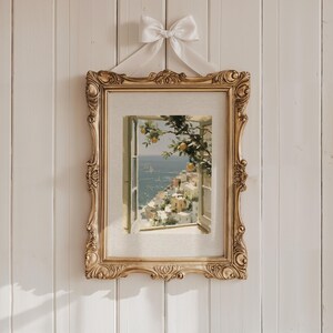 May include: A framed print of a coastal scene, featuring a window overlooking a town and the sea. The ornate gold frame is suspended by a white ribbon tied in a bow. The print depicts a sunny day with yellow citrus fruit.