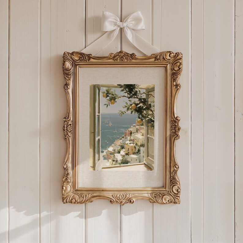 May include: A framed print of a coastal scene, featuring a window overlooking a town and the sea. The ornate gold frame is suspended by a white ribbon tied in a bow. The print depicts a sunny day with yellow citrus fruit.
