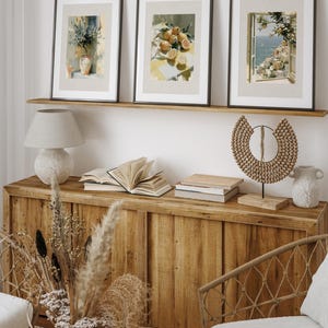 May include: Three framed art prints with black frames and beige mats are displayed above a wooden cabinet. The prints depict watercolor scenes of a vase with plants, lemons, and a coastal view. A lamp, books, and decorative items complete the scene.