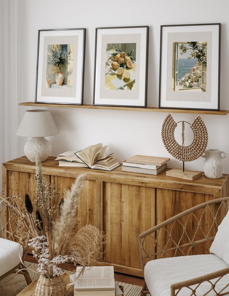 May include: Three framed art prints with black frames and beige mats are displayed above a wooden cabinet. The prints depict watercolor scenes of a vase with plants, lemons, and a coastal view. A lamp, books, and decorative items complete the scene.