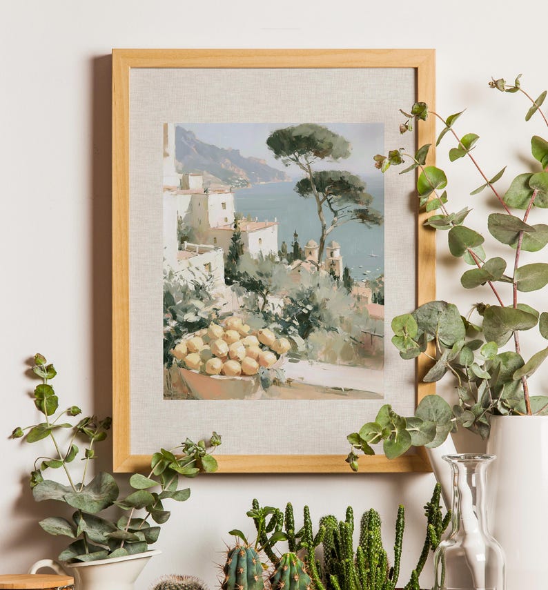 Amalfi Coast lemon terrace and umbrella pine Italian art print in a natural light wood frame mounted on a white wall surrounded by trailing eucalyptus, potted cacti, and succulent plants — loose impressionist oil-style painting of white Amalfi architecture, sea view and a bowl of lemons