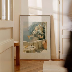 Large Positano hillside and balcony Italian coastal canvas art print in a natural wood float frame leaning against white French doors in a bright sunlit hallway — showing full canvas size with printed linen-textured mat design and warm golden light casting soft shadows across the white walls