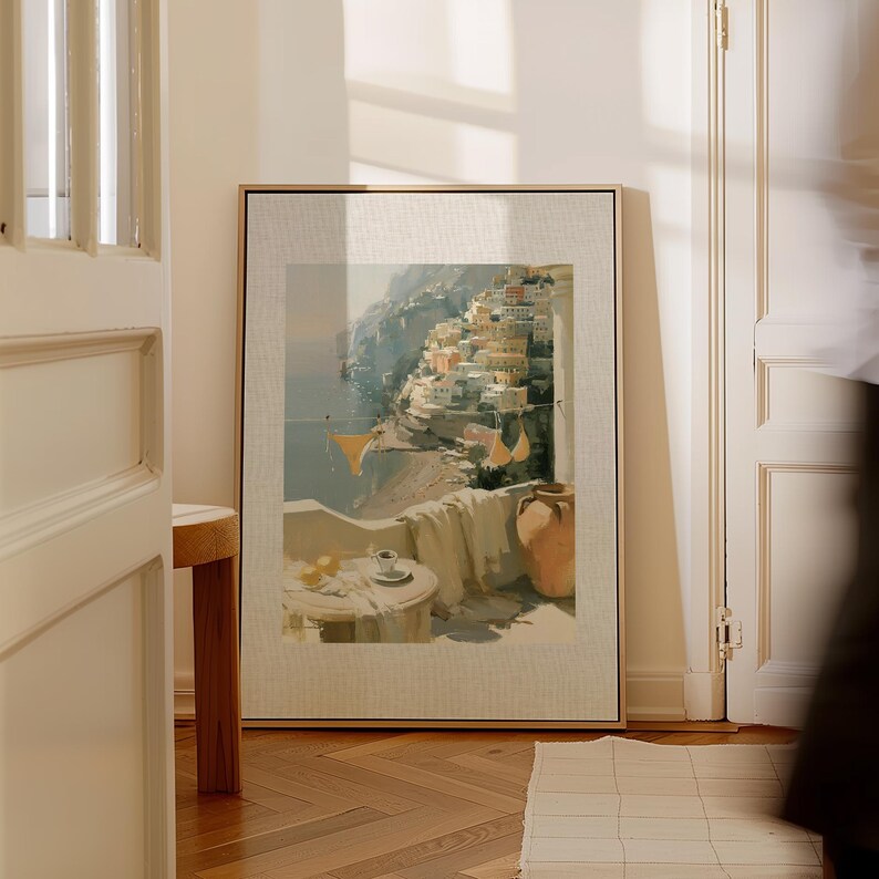 Large Positano hillside and balcony Italian coastal canvas art print in a natural wood float frame leaning against white French doors in a bright sunlit hallway — showing full canvas size with printed linen-textured mat design and warm golden light casting soft shadows across the white walls