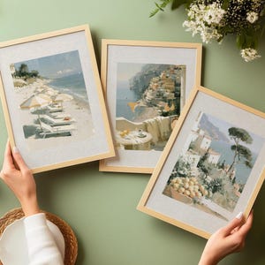 Hands holding three Amalfi Coast framed art prints fanned out on a sage green background styled with white spring blossom flowers and ceramic bowls — showing beach umbrella print, Positano balcony print, and Amalfi terrace lemon print in natural pine frames with printed linen mat