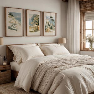 Three Amalfi Coast Italian art prints in dark walnut wood frames arranged as a triptych above a rustic wooden bed headboard in a warm cream bedroom — white linen bedding, knit throw blanket, bedside candle lanterns, natural linen curtains, and a wooden framed window with wildflowers