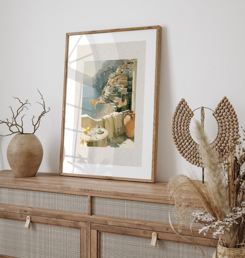 Positano hillside balcony Italian coastal art print in a dark oak wood frame leaning against a white wall on top of a rattan-door sideboard, styled with a round stone vase with bare branches, dried pampas grass, and a decorative seed pod sculpture — framed with printed linen-textured mat and a white mat border
