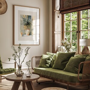 May include: A cozy living room scene featuring a framed watercolor painting of flowers in a vase. A green velvet sofa with matching pillows sits near a wooden coffee table with glass vases and floral arrangements. The room has a natural, earthy aesthetic.