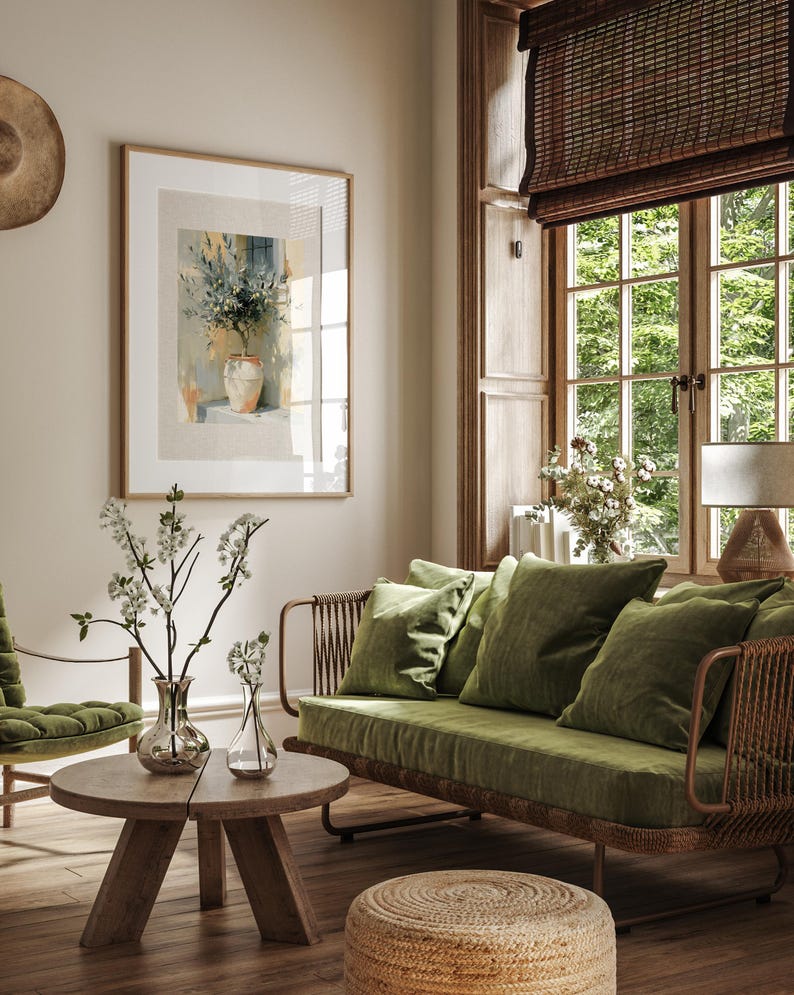 May include: A cozy living room scene featuring a framed watercolor painting of flowers in a vase. A green velvet sofa with matching pillows sits near a wooden coffee table with glass vases and floral arrangements. The room has a natural, earthy aesthetic.