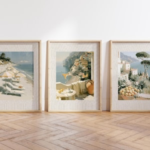 Set of 3 large Italian coastal oil-style art prints in wide natural wood linen-mat frames leaning against a white wall on herringbone light oak flooring — left print shows Amalfi beach with striped umbrellas, center shows Positano balcony terrace, right shows Amalfi lemon and umbrella pine view