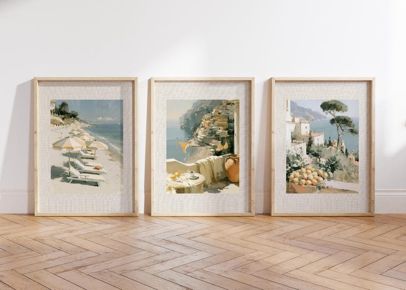 Set of 3 large Italian coastal oil-style art prints in wide natural wood linen-mat frames leaning against a white wall on herringbone light oak flooring — left print shows Amalfi beach with striped umbrellas, center shows Positano balcony terrace, right shows Amalfi lemon and umbrella pine view