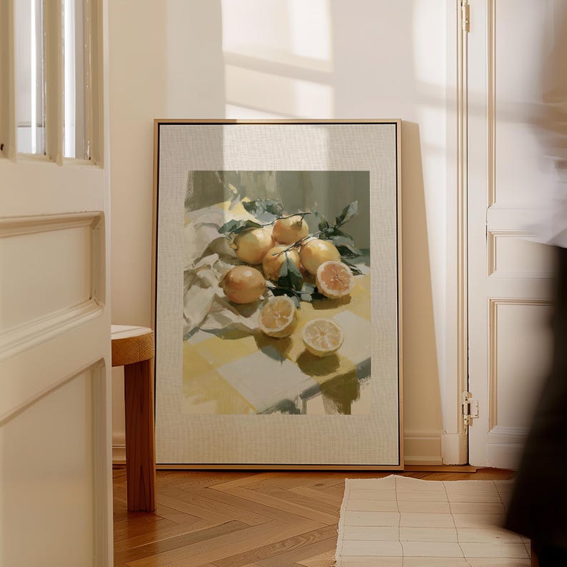 May include: A framed art print featuring a still life of lemons. The painting depicts whole and sliced lemons with green leaves on a yellow and white checkered surface. The artwork is displayed in a room with a wooden floor and a white door.
