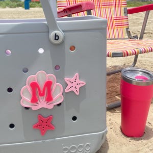 May include: A gray beach tote bag with seashell and starfish embellishments, a pink tumbler, and a striped beach chair on a sandy beach. The bag has a pink initial "M" on a seashell.