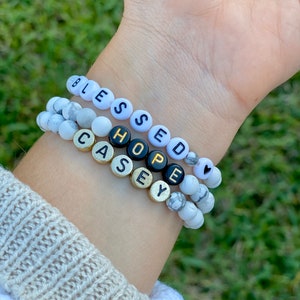 May include: Three bracelets with white marble beads and black letter beads spelling "Blessed", "Hope", and "Casey". The bracelets are stacked on a person's wrist.