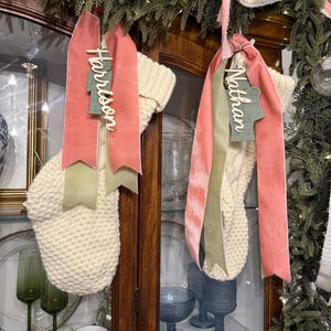 May include: Two cream-colored knit Christmas stockings hang from a wooden cabinet. Each stocking has a coral-colored ribbon and a green ribbon. One ribbon has the name "Harrison" and the other has the name "Nathan". The cabinet is decorated with greenery.