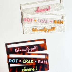 May include: Acrylic drink stirrers in two styles. One style is white with a pearlescent finish, and the other is tortoiseshell. Each stirrer has a rectangular shape with a slot. Text includes "cheers!", "DOT • CRAK • BAM", and "lets mahy yall!!".