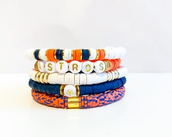Pode incluir: Um conjunto de cinco pulseiras com contas laranja, brancas e azuis. A pulseira superior tem contas brancas com letras douradas que soletram "ASTROS". A segunda pulseira tem contas douradas. A terceira pulseira tem contas brancas com uma estrela dourada. A quarta pulseira tem contas azuis. A pulseira inferior tem contas quadriculadas laranja e branco.