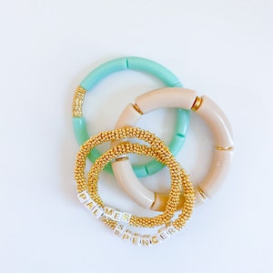 May include: Four bracelets stacked on top of each other. Two are light blue and two are beige. The beige bracelets have gold accents. The light blue bracelets have gold beads. The bracelets spell out the names "Palmer" and "Spencer".
