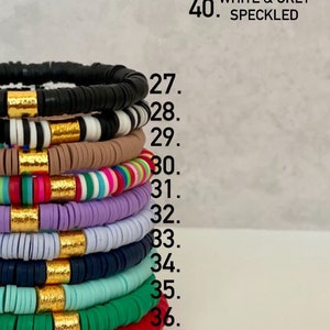 Puede incluir: Una pila de pulseras coloridas con detalles dorados. Las pulseras est&aacute;n hechas de cuentas redondas y est&aacute;n apiladas en una variedad de colores, incluyendo negro, blanco, marr&oacute;n, rosa, rojo, verde, azul y morado. El texto "NOT PICTURED // 38. RUST 39. CLEAR 40. WHITE & GREY SPECKLED" es visible sobre las pulseras.