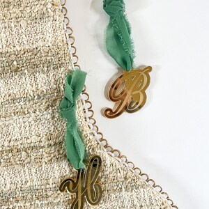 May include: Gold-colored acrylic letter ornaments, 'B' and 'H', suspended by green fabric ribbons. The letters are displayed against a textured, beige fabric with a scalloped edge. The overall aesthetic is elegant and decorative.