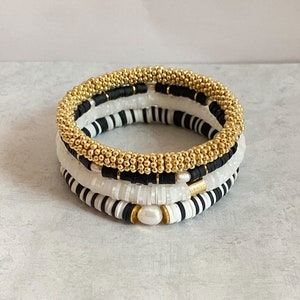 May include: A set of four bracelets featuring black and white beads and a gold beaded bracelet. The bracelets are stacked on top of each other and include a pearl accent.