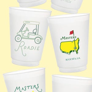 May include: White plastic cups with green and yellow golf-themed designs. The cups feature text and graphics related to the Masters Tournament, including the years 2026 and 1934, and the words "Roadie", "Augusta, GA", and "clubs, tees, & pimento cheese."