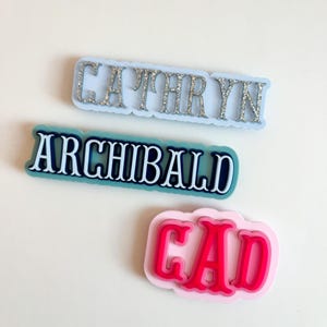 Puede incluir: Tres etiquetas con nombres en varios colores. La etiqueta superior es azul claro con letras plateadas con purpurina que deletrean "CATHRYN". La etiqueta del medio es verde azulado con letras blancas que deletrean "ARCHIBALD". La etiqueta inferior es rosa con letras rojas que deletrean "CAD".