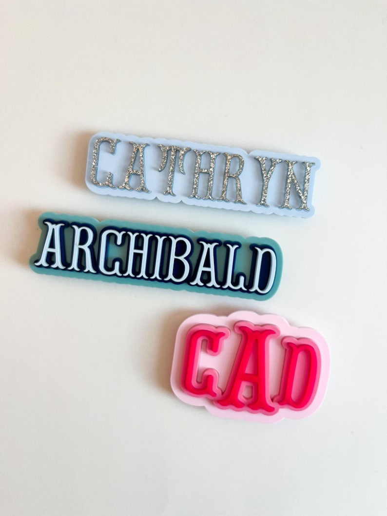 Puede incluir: Tres etiquetas con nombres en varios colores. La etiqueta superior es azul claro con letras plateadas con purpurina que deletrean "CATHRYN". La etiqueta del medio es verde azulado con letras blancas que deletrean "ARCHIBALD". La etiqueta inferior es rosa con letras rojas que deletrean "CAD".