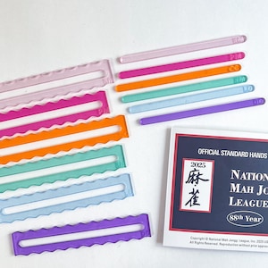 May include: A collection of colorful Mah Jongg game accessories, including clear plastic racks and matching pushers in various colors. A card with the text "OFFICIAL STANDARD HANDS" and "2025" is also present.