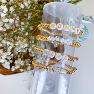 May include: Four gold beaded bracelets with white letter beads spelling out "MOMMY", "MAMA", "MAMA", and "MOM".