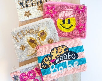 May include: Five beaded coin purses with different designs. The purses are pink, white, and turquoise with gold, black, and pink accents. The designs include the words "Yeehaw", "Cow Gal", "Rodeo Babe", a smiley face, and a cow.
